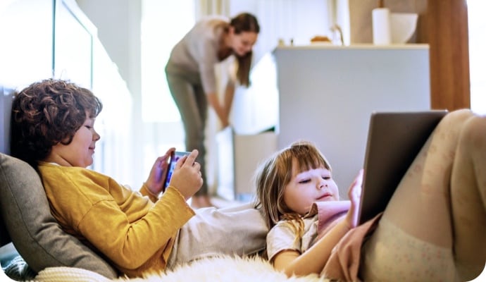 Kids lying down using tablet devices while mother in the background is tidying the kitchen.